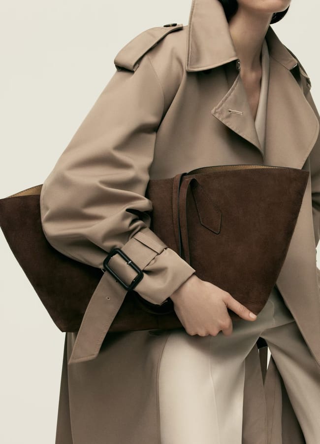 A person wearing a beige trench coat and white trousers holds a big brown suede clutch bag under their arm, the coat’s belt buckle visible and their face partially out of frame—a stylish take on the big bags trend.