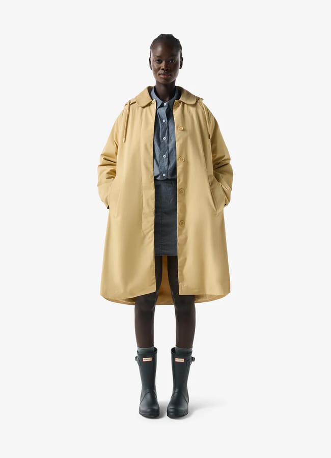A person stands facing forward in a beige trench coat—one of those stylish waterproof jackets—over a blue button-up shirt, grey skirt, and black wellington boots. Their hands are in the coat pockets against a plain white background.