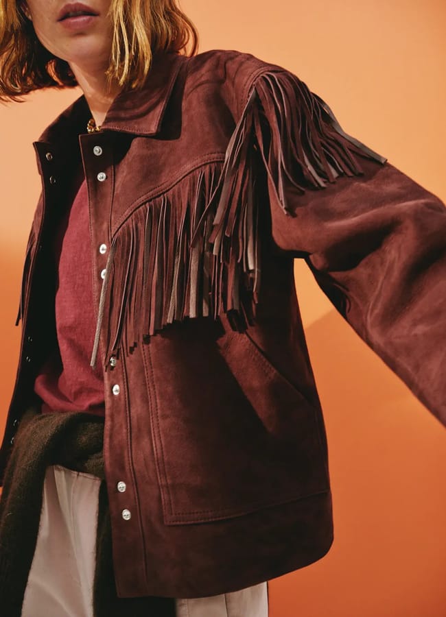 A person wearing a burgundy suede jacket with long fringe detailing—like stylish fringe benefits—over the chest and sleeves, paired with a maroon shirt and white trousers, poses in front of a peach-coloured background.