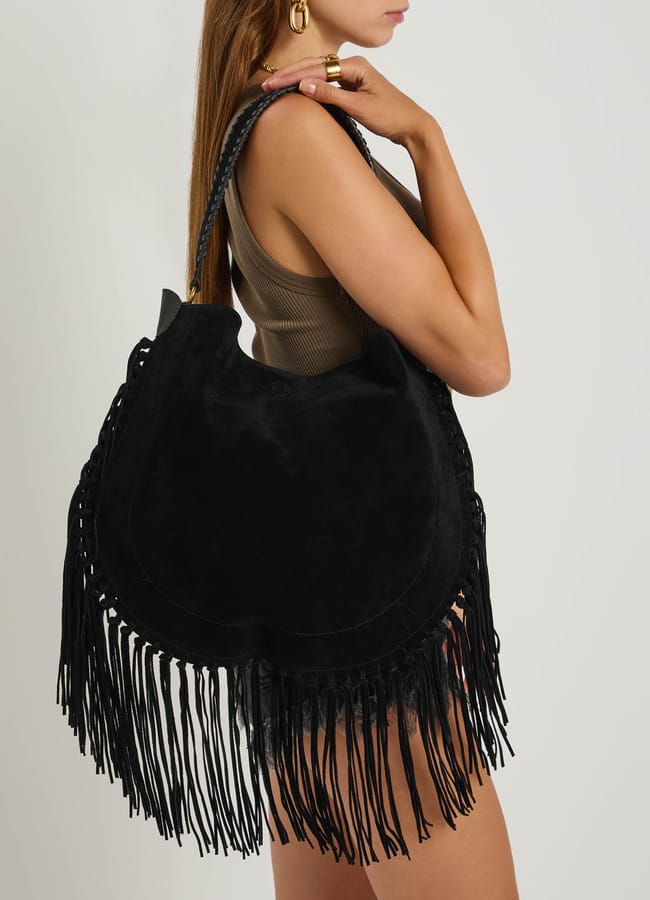 A woman in a sleeveless brown top and shorts holds a large black suede fringe handbag over her shoulder, showcasing stylish fringe benefits along the edges.