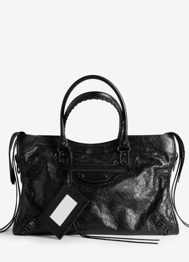 A black leather handbag with double handles, zip details, and a rectangular hanging tag, this stylish piece is perfect for those who love large bags. Displayed against a plain white background.