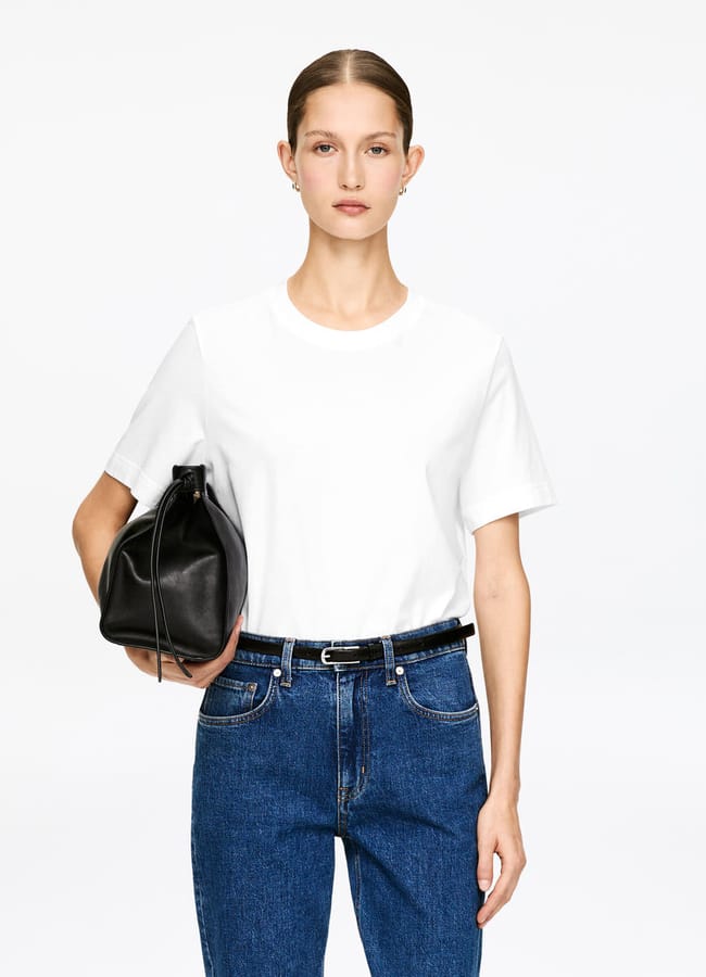 A person with straight brown hair pulled back wears a plain white T-shirt tucked into blue jeans, and holds a black handbag under their left arm, standing against a plain white background.