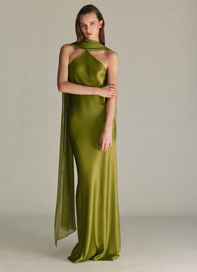 A woman stands in a studio wearing a floor-length, olive green satin gown with a halter neckline and draped scarf detail—an elegant choice for wedding guest dresses. She poses gracefully with straight brown hair and one arm crossed over her body.