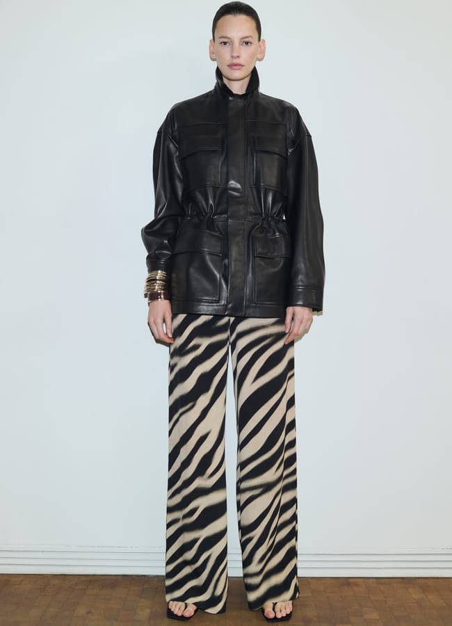 A person stands against a plain white wall wearing a black leather jacket, wide-legged zebra-print trousers, black sandals, and multiple gold bangles on their left wrist.