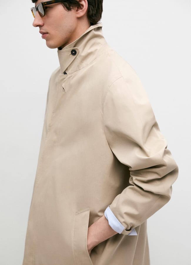 A person in a beige trench coat, one of this season’s must-have men’s jackets, stands in profile against a plain white background, wearing glasses and with one hand in their pocket.