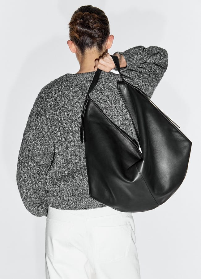 A person with hair in a bun, wearing a grey textured jumper and white trousers, is seen from behind holding one of this season’s must-have big bags—a large black leather slouchy bag—over their shoulder.