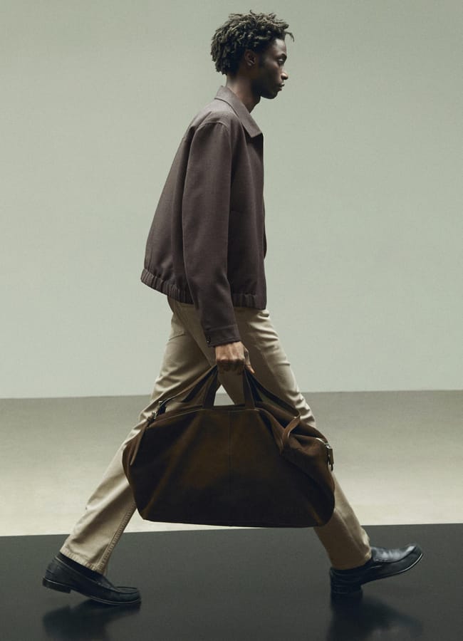 A person with short curly hair walks in profile, dressed in a brown men's jacket, beige trousers, and black shoes, carrying a large brown bag by their side against a minimalist background.