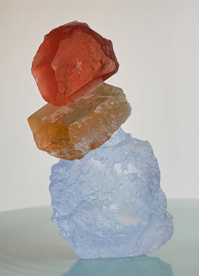 Three translucent crystals—red, amber, and blue—are stacked vertically on a smooth, light-coloured surface against a plain background, evoking the vibrant design sensibility admired by Interiors Insider and Lucy Barlow of Barlow & Barlow.