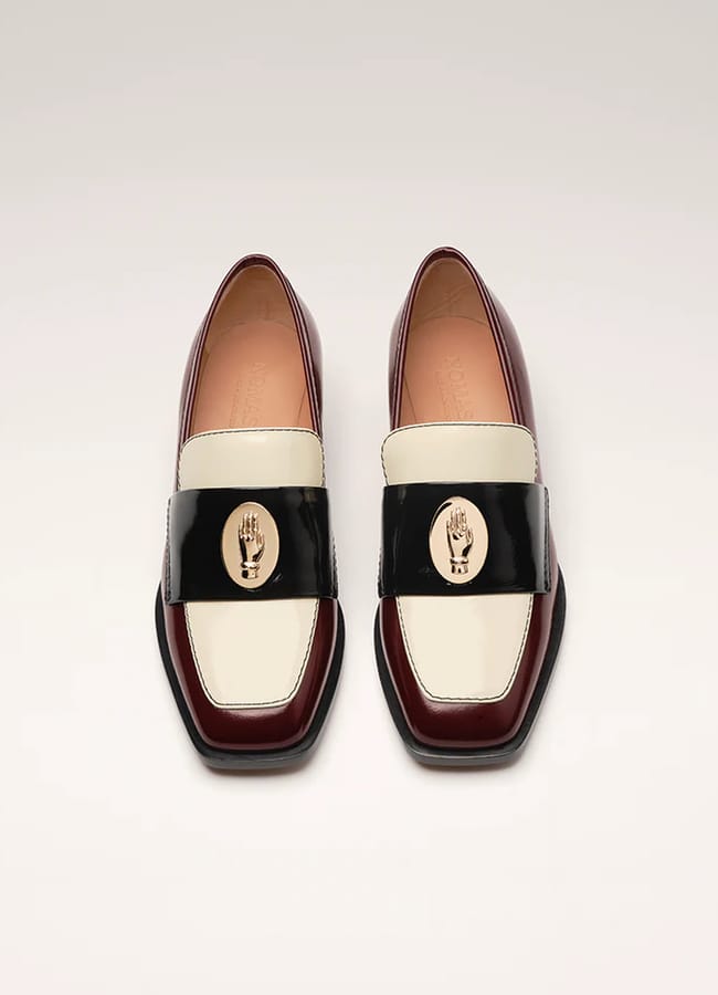 A pair of burgundy, cream, and black leather loafers with square toes and gold oval emblems featuring an engraved hand design on the front bands—an elegant nod to spring summer 2026 fashion trends.