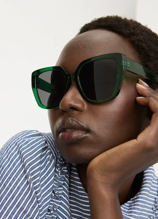 A person wearing oversized, dark green sunglasses and a blue and white striped top rests their head on their hand, gazing slightly upwards.
