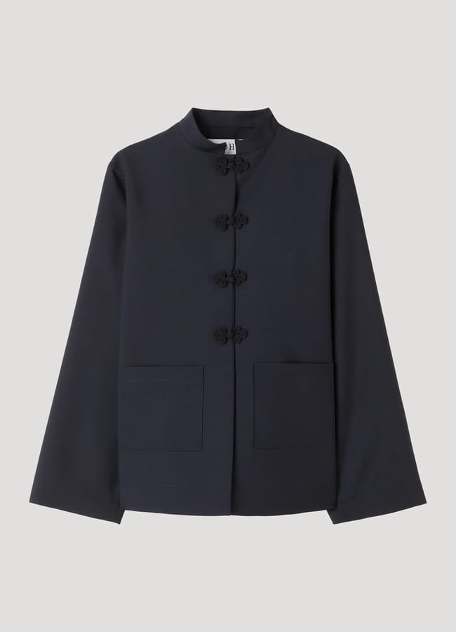 A navy blue, long-sleeve jacket with a mandarin collar, decorative knotted button fastenings down the front, and two large front pockets—perfectly embodying spring/summer 2026 fashion trends—displayed on a plain light grey background.