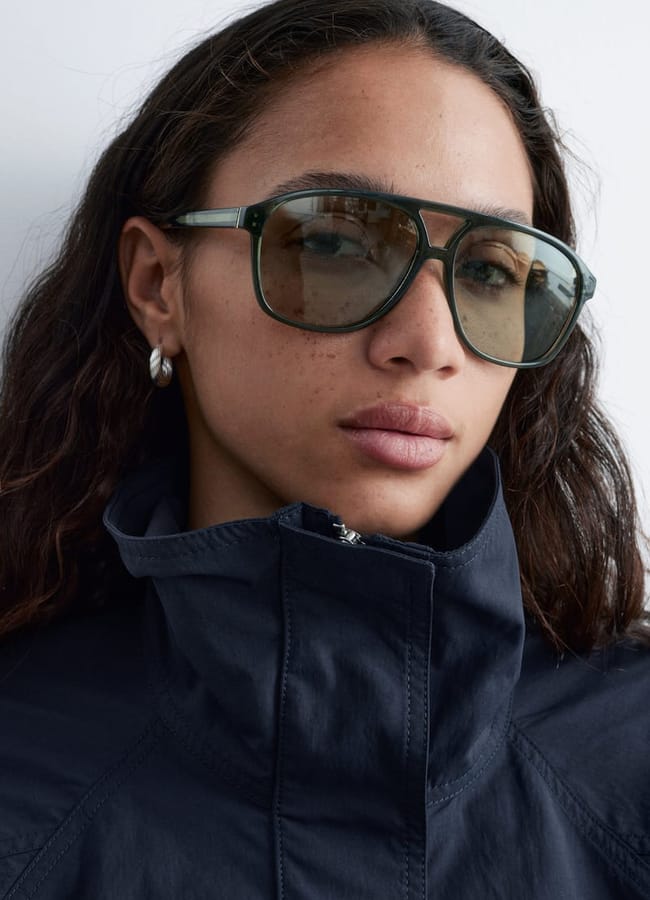 A person with long curly hair wears large, square sunglasses, silver hoop earrings, and a dark zip-up jacket with the collar up, standing in front of a plain white background.