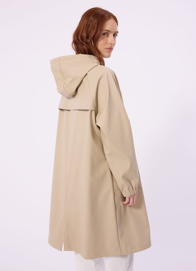 A person with long red hair wears a stylish waterproof jacket—a beige hooded raincoat with an overlapping back flap—standing sideways and looking over their shoulder against a plain light background.