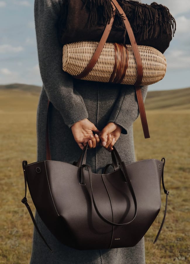 A person in a long grey coat stands outdoors, holding a large dark brown leather tote bag—perfect for fans of big bags. They also carry a rolled-up brown blanket and a wicker basket tied with brown leather straps, with grass and hills in the background.