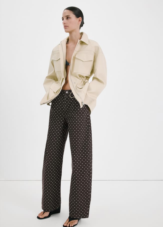 A woman stands against a white background, wearing a beige oversized jacket, a bikini top, wide-leg brown trousers with white polka dots, and black sandals. Her hands are in her pockets and her hair is slicked back.