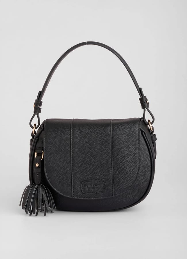 A black leather handbag with a curved flap, single top handle, gold fittings, and a tassel accent on one side, displayed against a sheer light grey background.