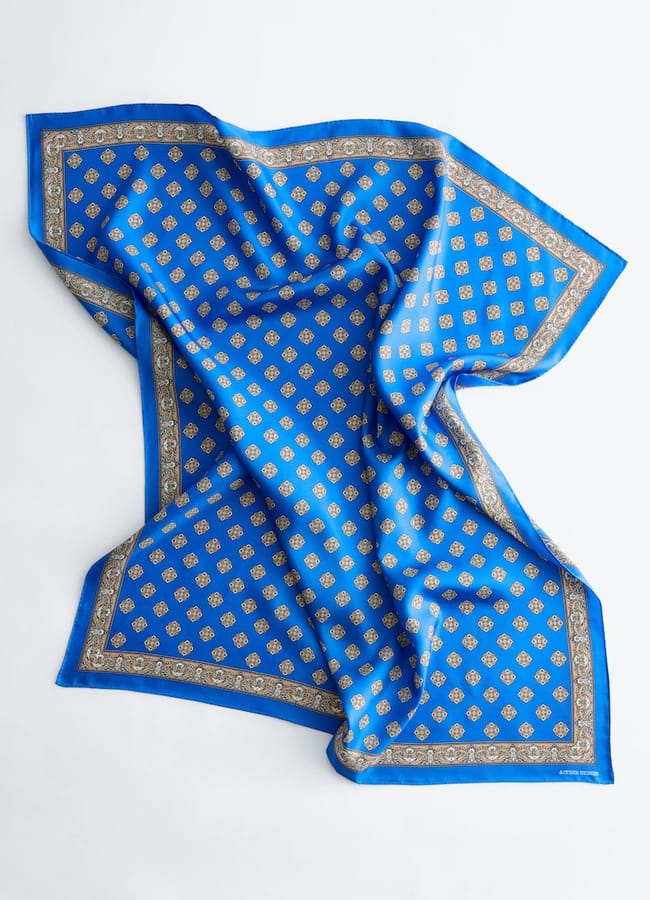 A square blue silk scarf with intricate gold geometric patterns and a decorative gold border, laid flat on a light surface with soft folds and creases—an elegant accessory inspired by SS26 trends.