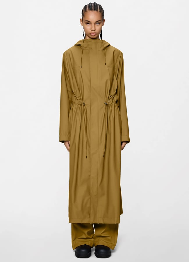 A person stands facing forwards, wearing a stylish waterproof jacket in olive-green with a hood and drawstring waist, matching trousers, and black shoes against a plain white background.
