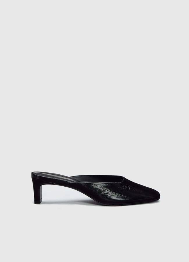A black, pointed-toe mule shoe with a low heel and a sheer accent is positioned against a plain light grey background.