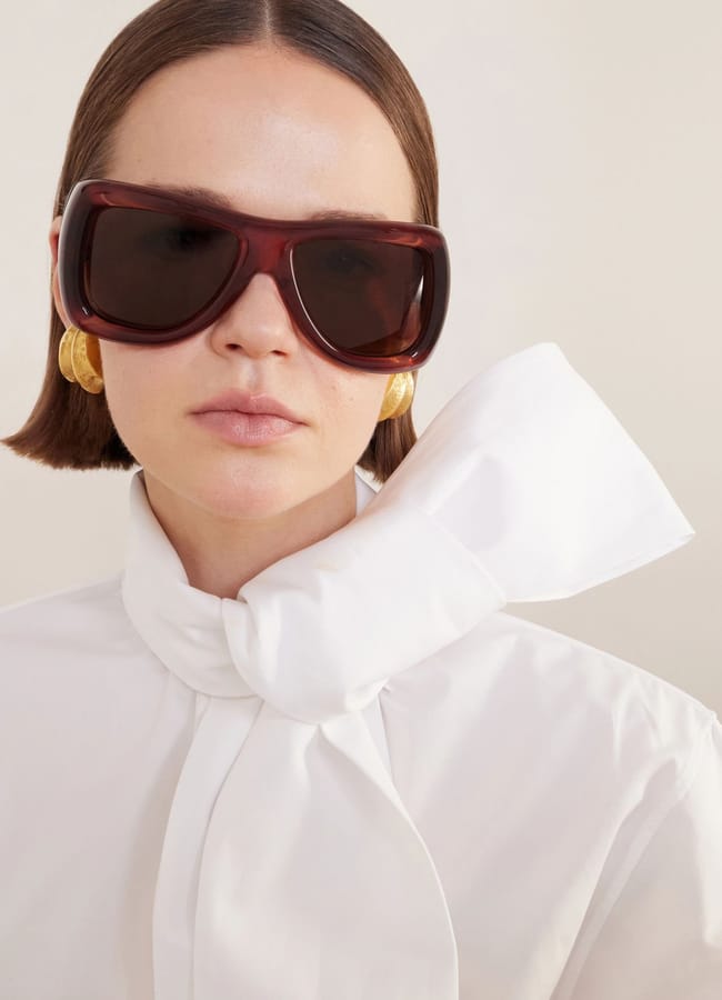 A woman with straight brown hair wears oversized dark sunglasses, large gold earrings, and a white blouse with a large bow at the neck, standing against a light background.