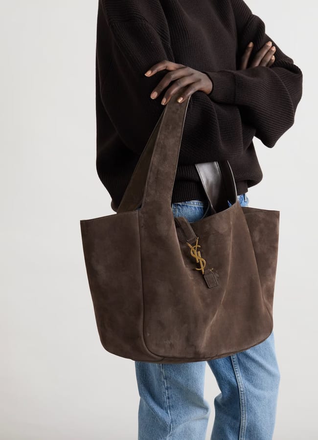 A person wearing a dark jumper and light blue jeans carries a large brown suede shoulder bag with a gold YSL logo and leather details, standing against a plain background—perfect for those who love big bags.