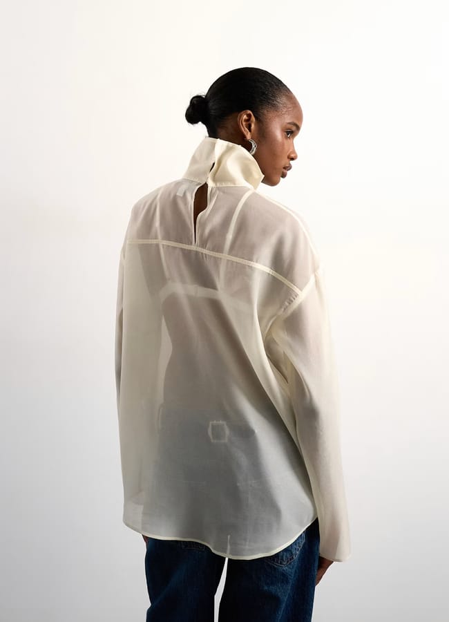 A woman with her hair in a bun faces away, wearing a sheer, off-white long-sleeve blouse with a high collar and navy jeans, set against a plain white background.