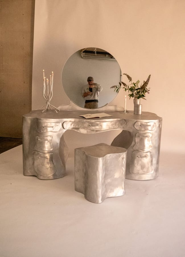 A shiny, sculptural silver dressing table by Barlow & Barlow with a matching stool, a round mirror, a candelabrum with lit candles, and a vase of flowers sits against a neutral background, while a person is reflected in the mirror.