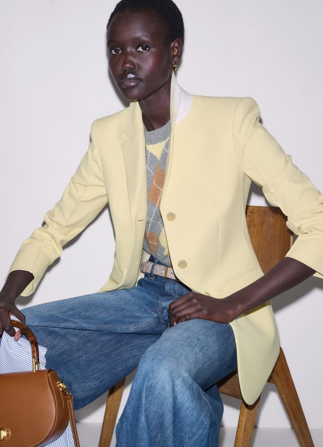 A woman sits on a wooden chair against a plain background, channelling her inner Style Editor in a pale yellow blazer, argyle jumper, blue jeans, and holding one chic brown handbag as her statement piece.