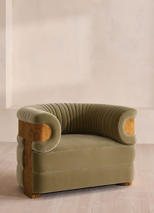 A green velvet armchair with rounded, ribbed backrest and wooden accents adds a touch of green decor, standing on a light wooden floor against a beige textured wall.