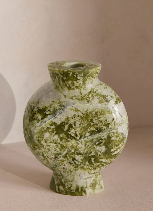 A round, marble vase with a narrow opening and green-and-white marbled pattern serves as striking green decor atop a beige surface, casting a soft shadow against a light, textured wall.