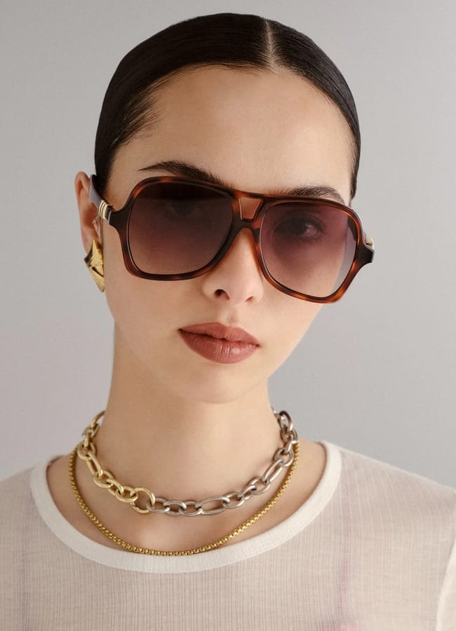 Woman with slicked-back dark hair wearing oversized brown sunglasses, gold and silver chain necklaces, a gold earring, and a sheer white top, posing against a plain light background.