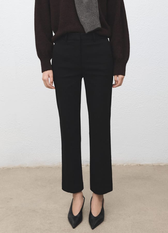 Channelling Carolyn Bessette Kennedy’s cool fashion, a person in black straight-leg trousers, a dark long-sleeved top, and pointed-toe shoes stands against a plain light wall. Their head is not visible in this '90s wardrobe-inspired image.