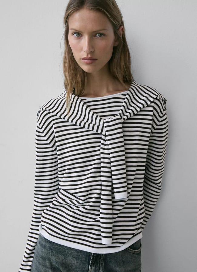 Woman standing against a white wall, wearing a black and white striped jumper with a matching jumper draped over her shoulders—an effortlessly chic look that captures spring summer 2026 fashion trends. She pairs it with dark jeans and wears her light brown hair straight.