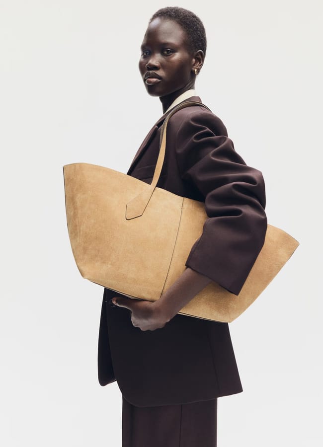 A woman in a dark brown suit poses against a plain light background, holding one of this season's big bags—a large tan suede tote—under her arm.