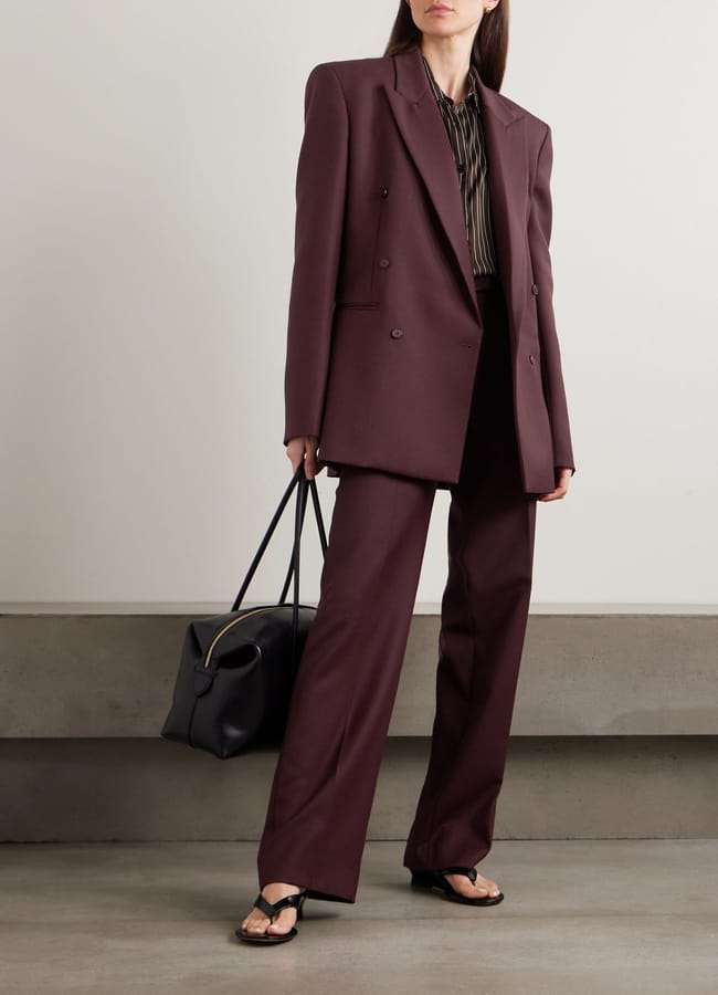 A woman stands indoors wearing a loose-fitting burgundy suit—a true fashion essential—paired with a striped blouse, black sandals, and a large black handbag. Her hair is straight and loose, and her face is partially out of frame.
