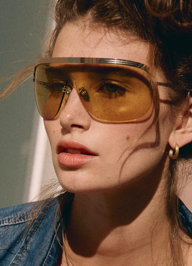 A woman with wavy brown hair wears oversized yellow-tinted sunglasses, gold hoop earrings, and a blue denim jacket. She gazes slightly to the side with a neutral expression in soft natural light.