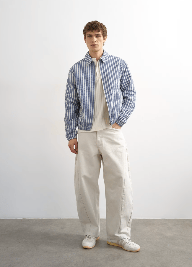 A young man stands against a plain background wearing a blue and white striped men's jacket over a cream shirt, loose-fitting beige trousers, and white trainers. He has short curly hair and a neutral expression.