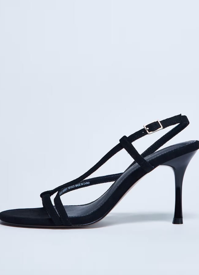 A black heeled sandal with a thin stiletto heel, open toe, and minimalist sheer straps, including an ankle strap with a small buckle. The sandal is displayed on a plain light background.
