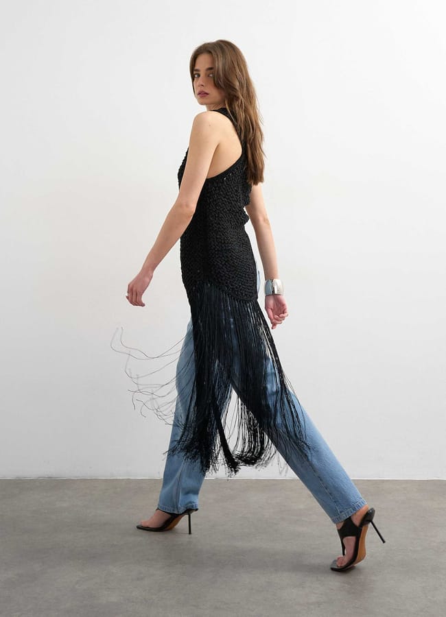 A woman in a sleeveless black top with long fringe, light blue jeans, and black high heels walks against a plain white wall; she has long brown hair and wears a silver cuff bracelet—her style echoes fashion’s take on fringe benefits.