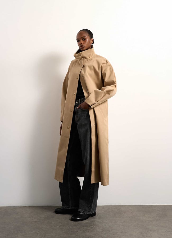 A person stands against a plain white wall wearing a long beige trench coat over a black top and wide-leg dark jeans, with black shoes. The coat’s collar is turned up and one hand is in a pocket.