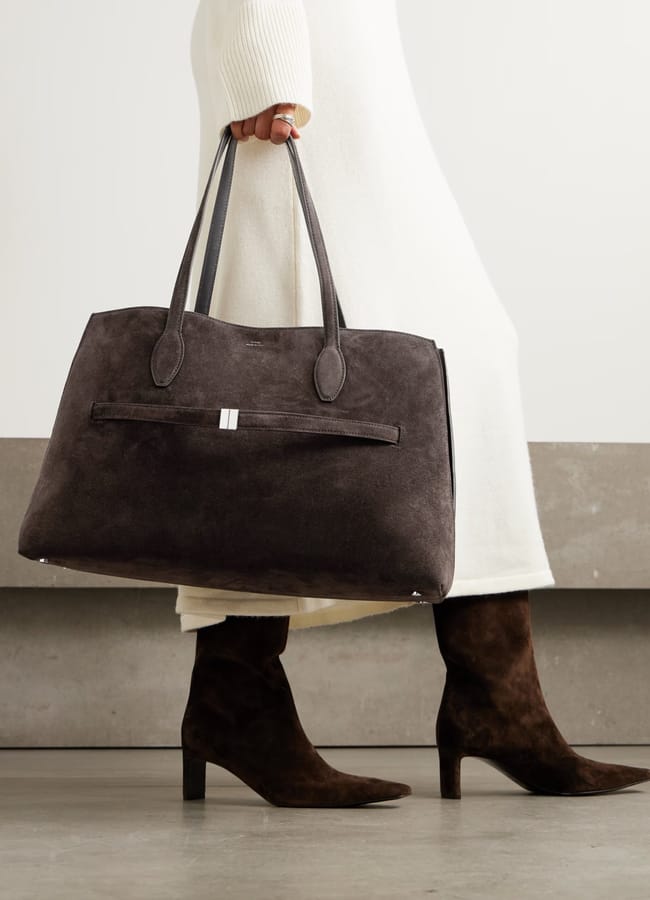 A person in a white dress and dark brown suede boots carries one of this season's big bags—a large dark brown suede tote with long handles—walking on a smooth floor against a light background.