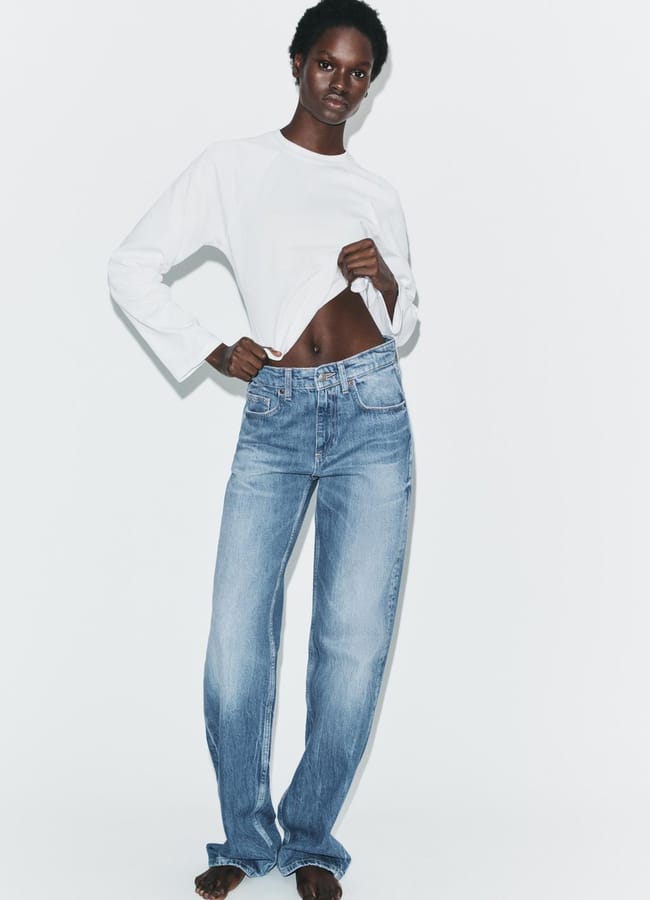 A person stands barefoot against a white background, wearing a white long-sleeved shirt and light blue straight-leg jeans, with one hand lifting the shirt slightly to reveal their midriff.