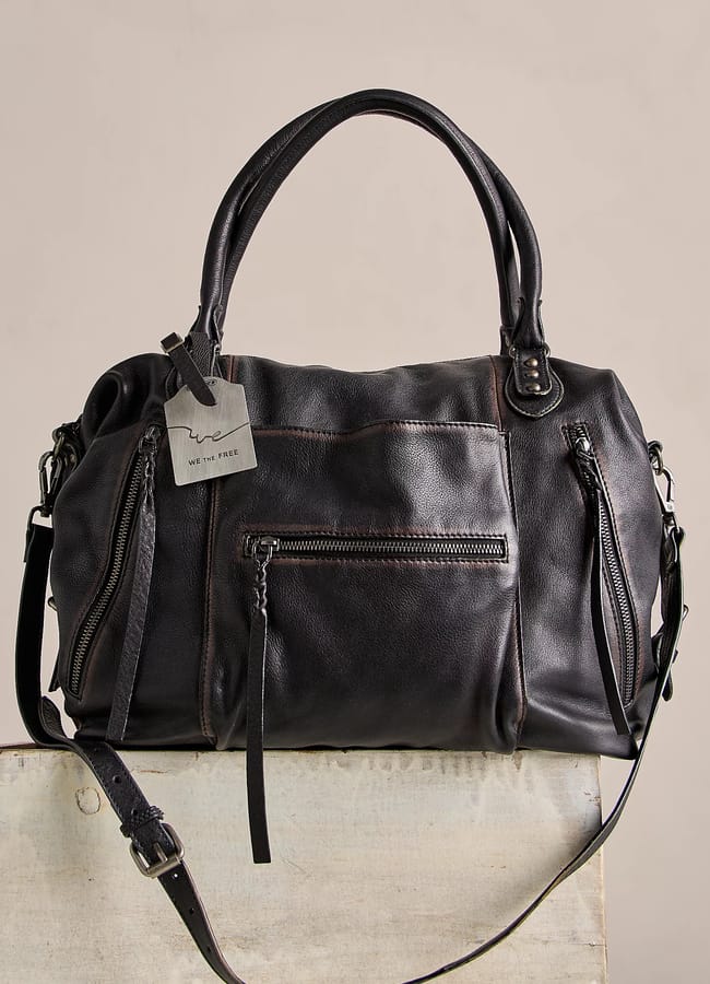 A black leather handbag, perfect for those who love big bags, with two handles, a detachable shoulder strap, and multiple zipped pockets, displayed on a light wooden surface with a small grey tag attached to one handle.