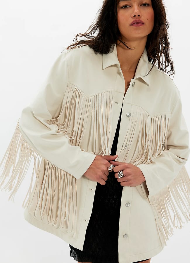 A woman wearing a light-coloured jacket with long fringe benefits details across the chest and sleeves over a black dress, standing against a plain white background.