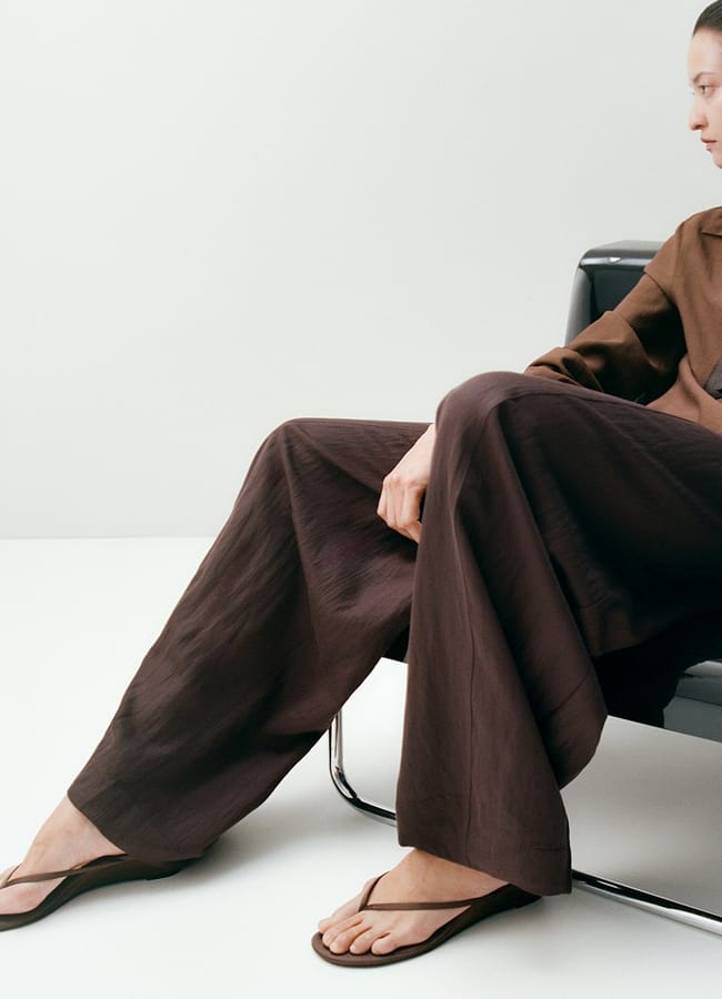 A person wearing a brown shirt, loose brown trousers, and black flip-flops sits on a black chair against a plain white background. Only their upper body and legs are visible.