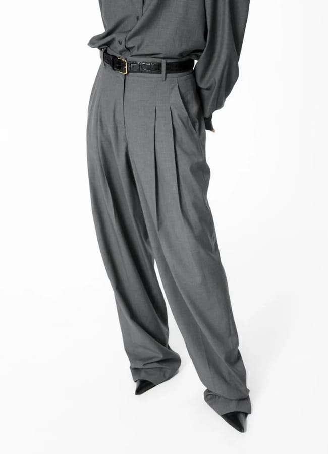 Person wearing loose-fitting, high-waisted grey dress trousers with pleats, a black belt, and a matching grey shirt; their hands are in their pockets and pointed black shoes complete this effortlessly chic wardrobe look you can easily recreate.
