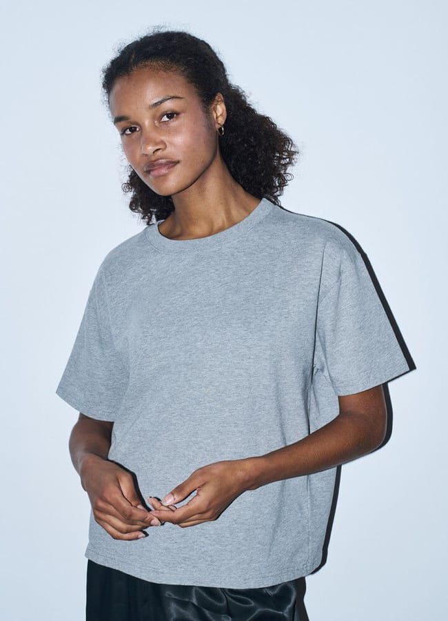 A woman with curly hair stands against a plain light background, wearing a loose-fitting grey T-shirt and dark trousers, looking calmly at the camera with her hands relaxed in front of her.