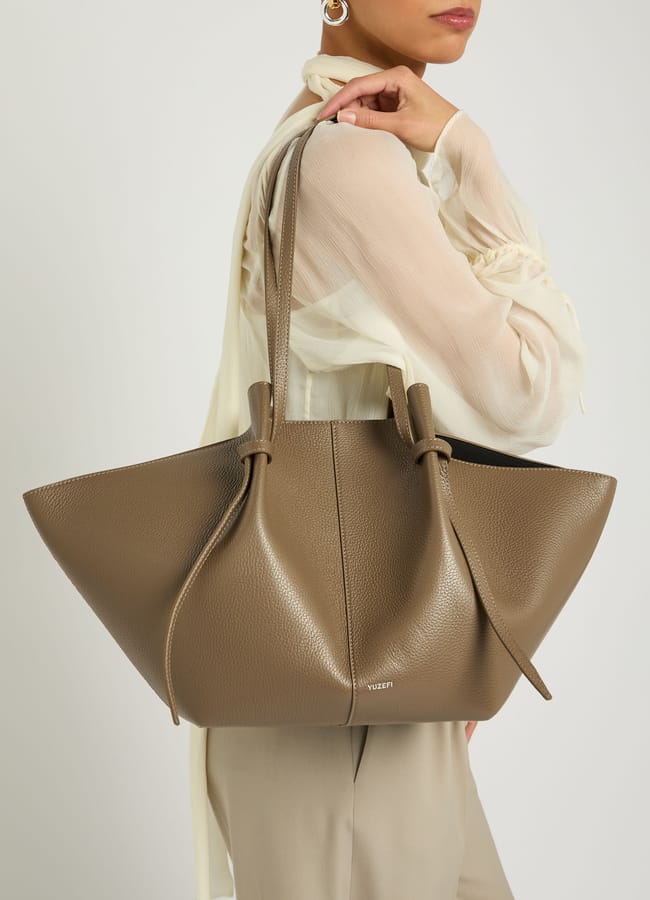 A woman wearing a sheer, long-sleeved blouse and gold hoop earrings carries a stylish big bag—a large taupe leather tote with knotted straps—over her shoulder.