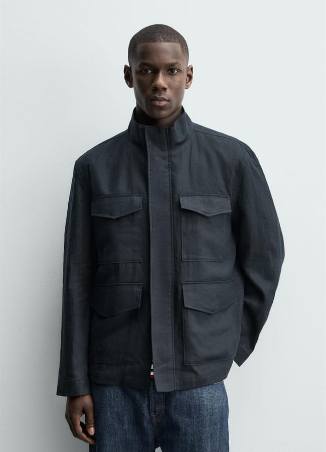 A person wearing a dark, collared men's utility jacket with large front pockets and blue jeans stands against a plain light-coloured background.