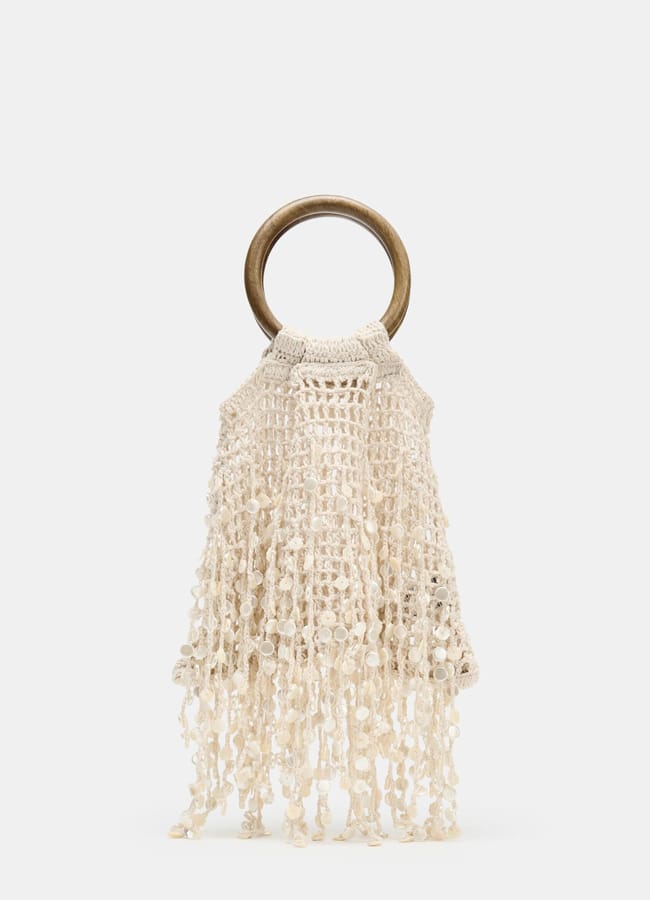 A sheer macramé handbag with a large round wooden handle and an off-white, fringed, netted design, displayed against a plain white background.